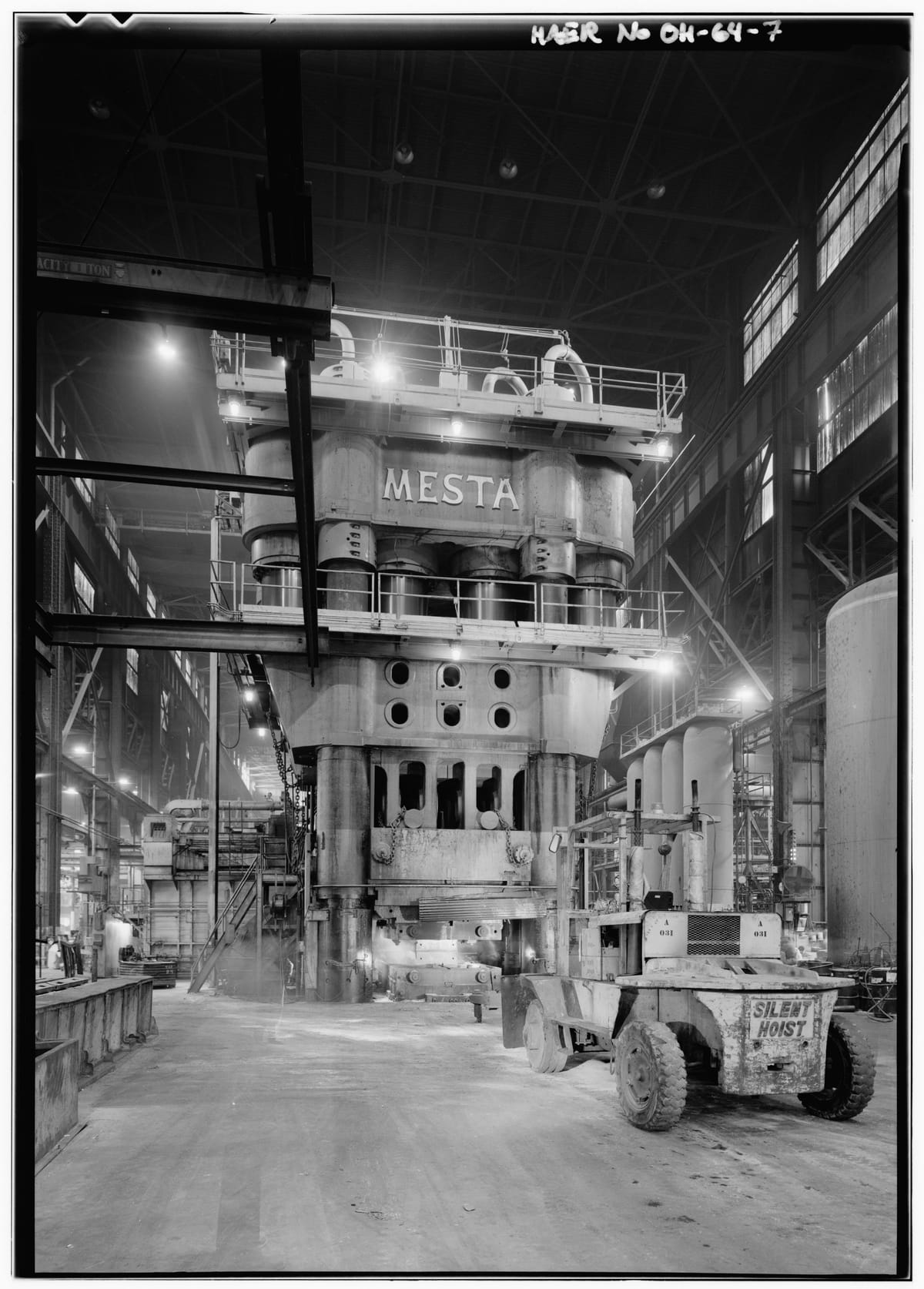 A tall press in a lit forge hall, its top crosshead lettered MESTA above a row of cylinders and a railed platform, with a forklift truck in the foreground.