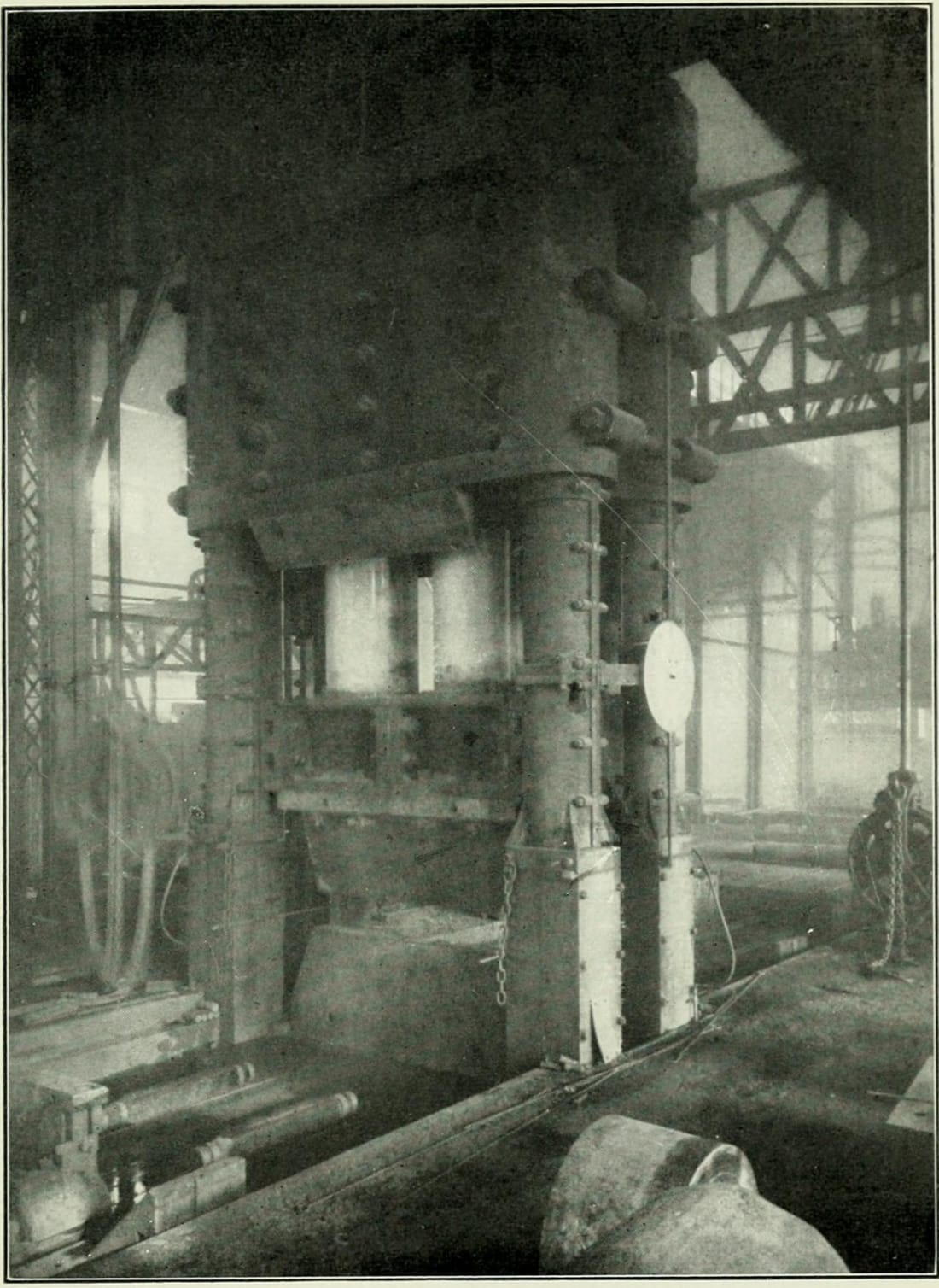 A tall hydraulic forging press in a steel-framed shop: heavy columns with large nuts, the crosshead above a block on the anvil, a pressure gauge on one column, and a large round forging on the floor in front.