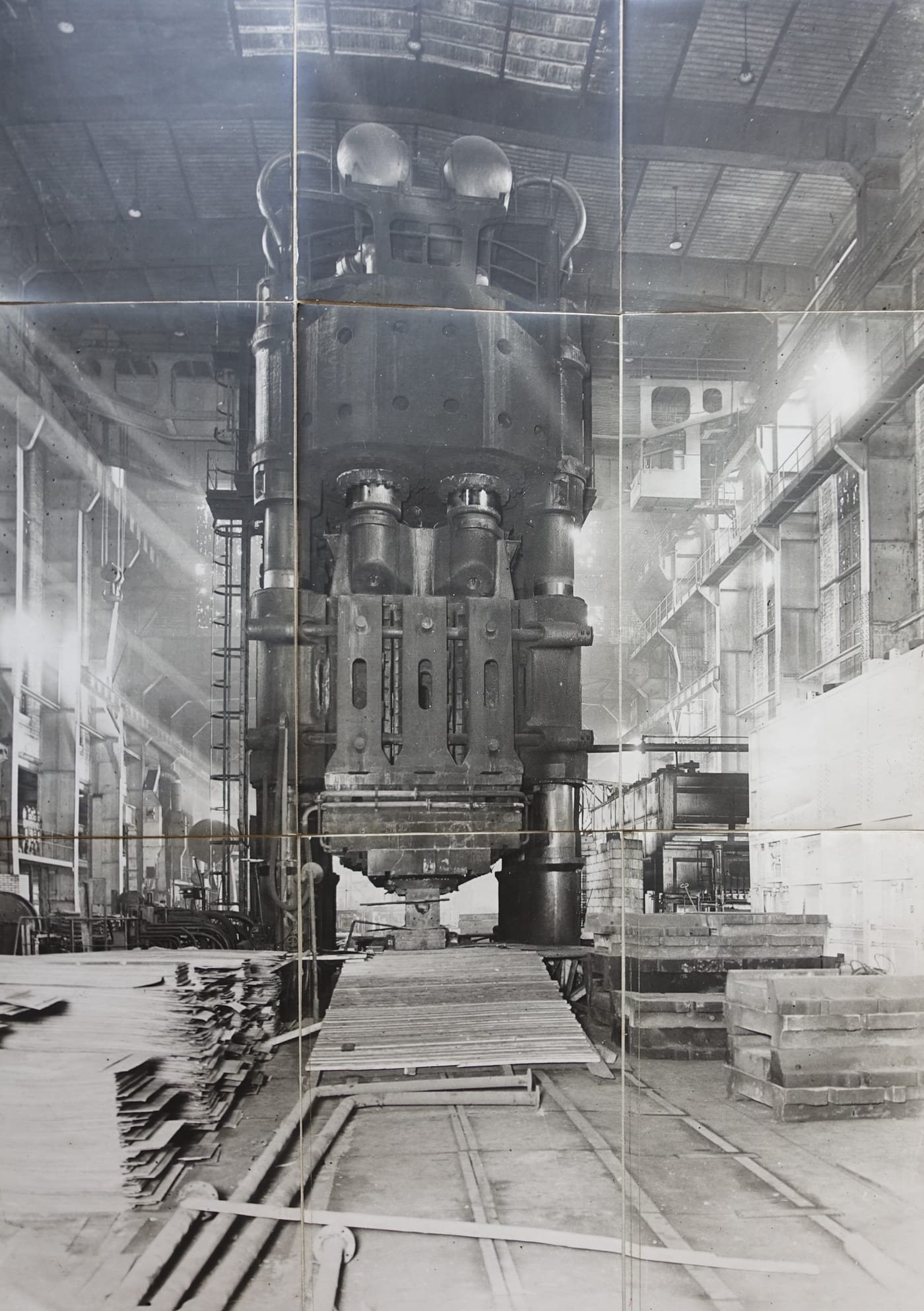 Black-and-white photograph of a very tall press seen end-on in a high factory hall: two domed tanks and curved pipes on top, a massive crosshead, two rams, a slotted moving crosshead between the columns, a ladder up the left column, and floor planks and rails in front.