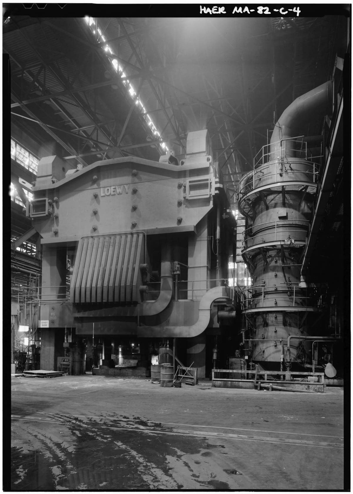 A massive press in a dark forge hall: a broad steel housing lettered LOEWY above a ribbed guard, with a tall tower of pipes and platforms beside it.